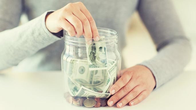 A person adds a dollar to a jar of saved money.
