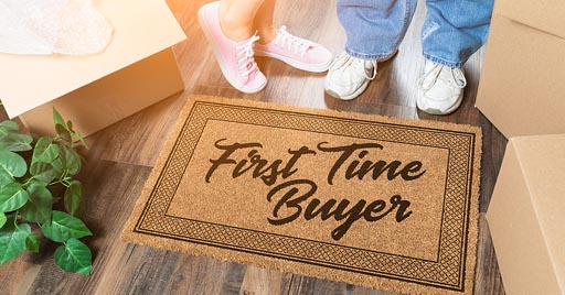 A couple stands next to moving boxes and a front door mat reads First Time Home Buyer.